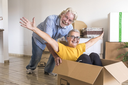 A senior woman sitting in a moving box with arms stretched out and smiling and a senior man pushing the box and smiling