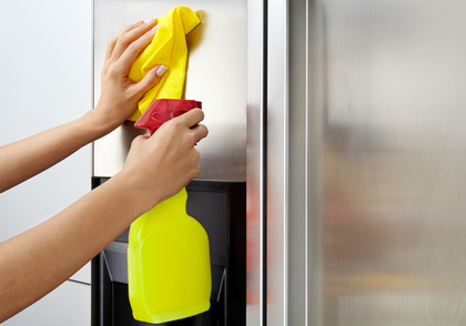 Left hand wiping a stainless steel fridge with a yellow cloth and right hand spraying it with a yellow spray bottle